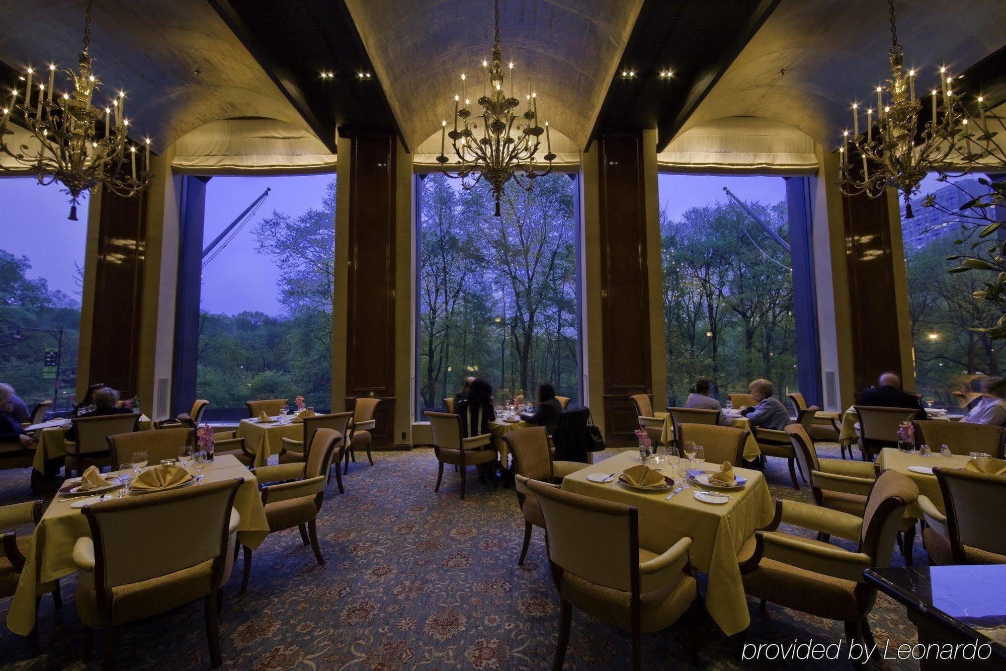 Park Lane New York Hotel Restaurant photo The photo depicts an elegant dining setting within a restaurant. The space features several tables, each set with white tablecloths and neatly arranged dining accessories. There are opulent chandeliers hanging from the ceiling that provide a warm glo